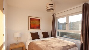 Ein Schlafzimmer mit einem großen Bett, einem Nachttisch mit Lampe, einem Bild an der Wand und einem Fenster mit Vorhängen.