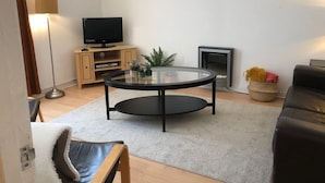 A living room with a round glass table, a flat-screen TV on a wooden cabinet, a fireplace, and a black leather sofa. - Lovely one-bedroom apartment just a few yards off Edinburgh’s famous Royal Mile. Sleeps 2. (Edinburgh)