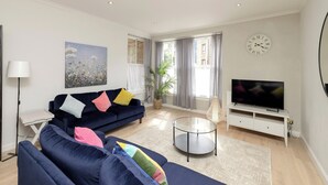 A modern living room with a dark blue sofa, a glass coffee table, a flat-screen TV, and a large window with curtains.