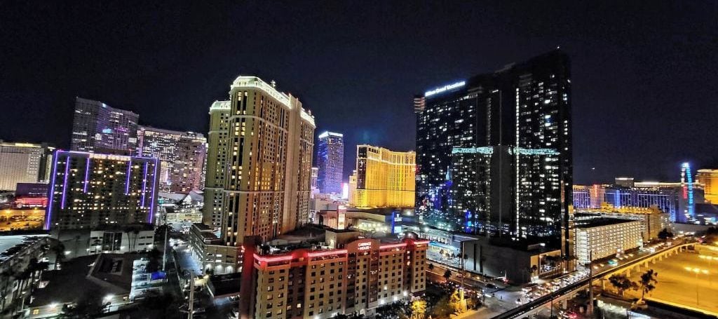 A nighttime cityscape with illuminated buildings, including a prominent hotel with a classic design and a modern skyscraper with lit windows.