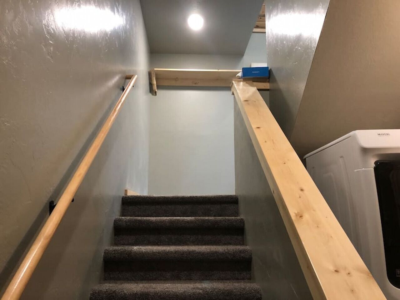 Un escalier avec des marches recouvertes de moquette et des rampes en bois, menant à un palier où se trouvent un radiateur et un objet bleu accroché au mur.
