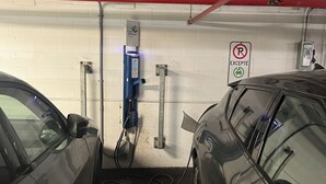 Un estacionamiento con dos coches aparcados, un cartel de prohibido estacionar con una excepción y un cuadro eléctrico en la pared.