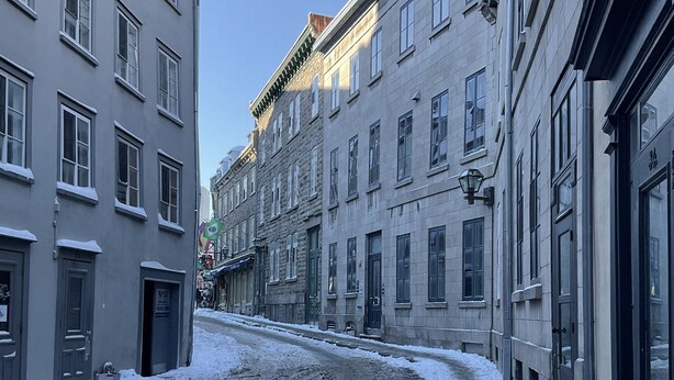 Una calle cubierta de nieve bordeada por edificios de varios pisos con ventanas y puertas amplias, un cielo despejado y un farol a la vista.