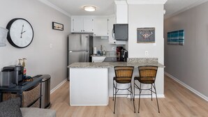 A compact kitchen with white cabinets, a stainless steel refrigerator, a built-in oven, and a small island with two wicker bar stools. - The Lookout: Tranquil Beachfront Condo (North Topsail Beach)