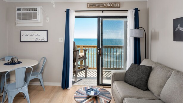 A room with a balcony overlooking the sea, a dining table with chairs, a sofa, and a sign reading "THE LOOKOUT". - The Lookout: Tranquil Beachfront Condo (North Topsail Beach)