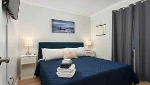 A neatly made bed with a blue quilt, two bedside lamps, a stack of folded towels, a framed picture of a lighthouse, and a grey curtain. - The Lookout: Tranquil Beachfront Condo (North Topsail Beach)
