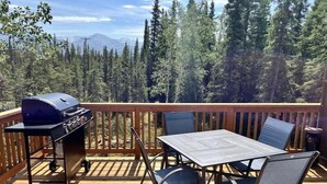 A deck with a barbecue grill, a table, and chairs, overlooking a forested area with mountains in the distance.