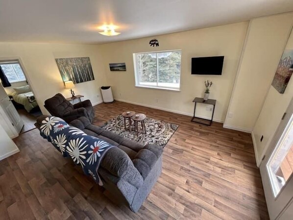 A living room with a gray sofa, a patterned throw blanket, a small rug, and a flat-screen TV mounted on the wall.