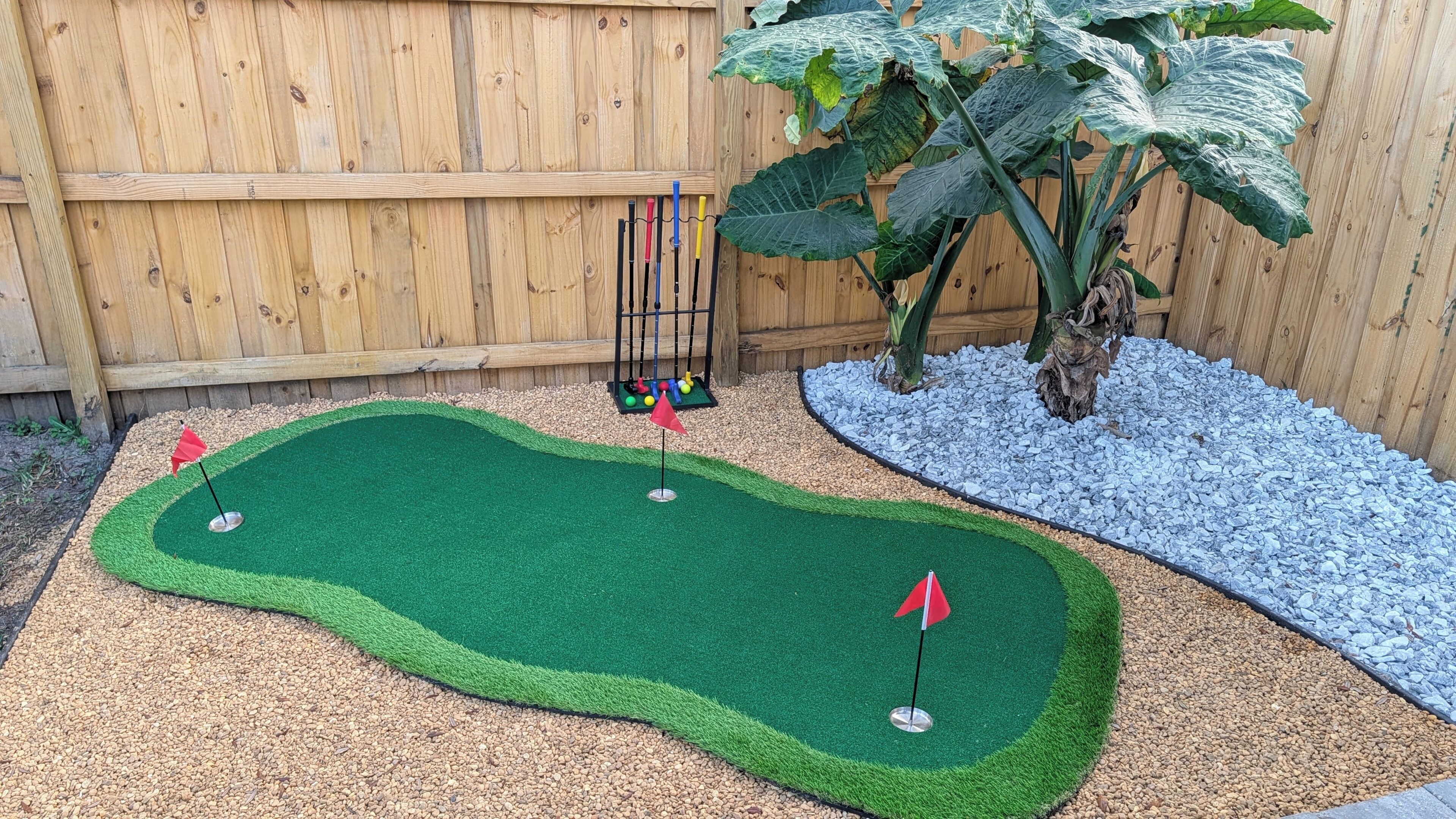 A miniature golf course with a wooden fence, a large plant, and a pebbled area.