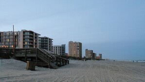 Een strand met een houten trap die naar een gebouw leidt, zandstrand en hoge gebouwen op de achtergrond.