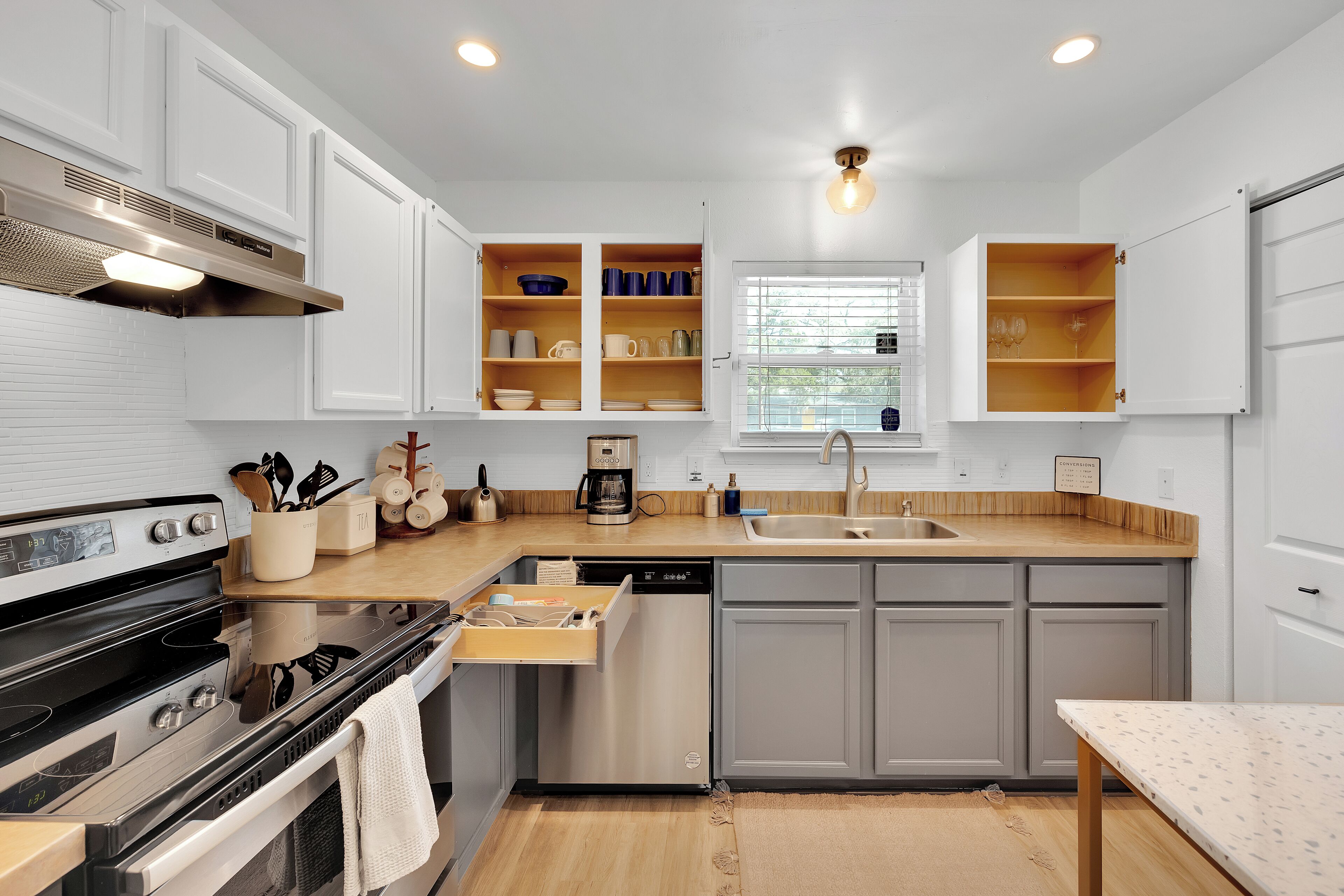Une cuisine moderne avec des meubles blancs, une cuisinière en acier inoxydable, un lave-vaisselle et une fenêtre avec des stores.