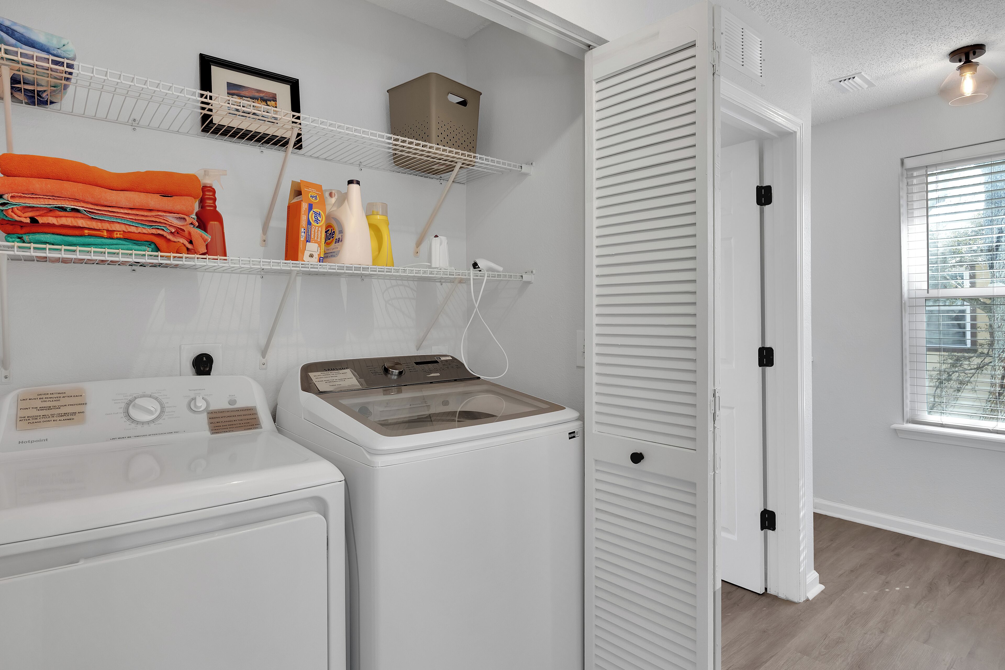 A laundry area with a front-loading washer and dryer, shelves with folded clothes and laundry supplies, and a window with blinds.