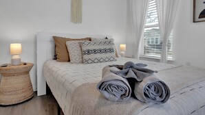 Een net opgemaakt bed met opgerolde handdoeken, een bedlampje en een gestreept kussen op een wit hoofdbord.