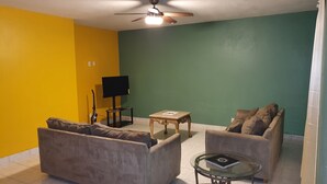 Uma sala de estar com dois sofás, uma mesa de centro de vidro, uma televisão sobre um suporte e um ventilador de teto.
