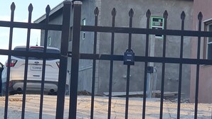 Uma área de estacionamento cercada, com piso de concreto, uma cerca de metal e um edifício ao fundo.
