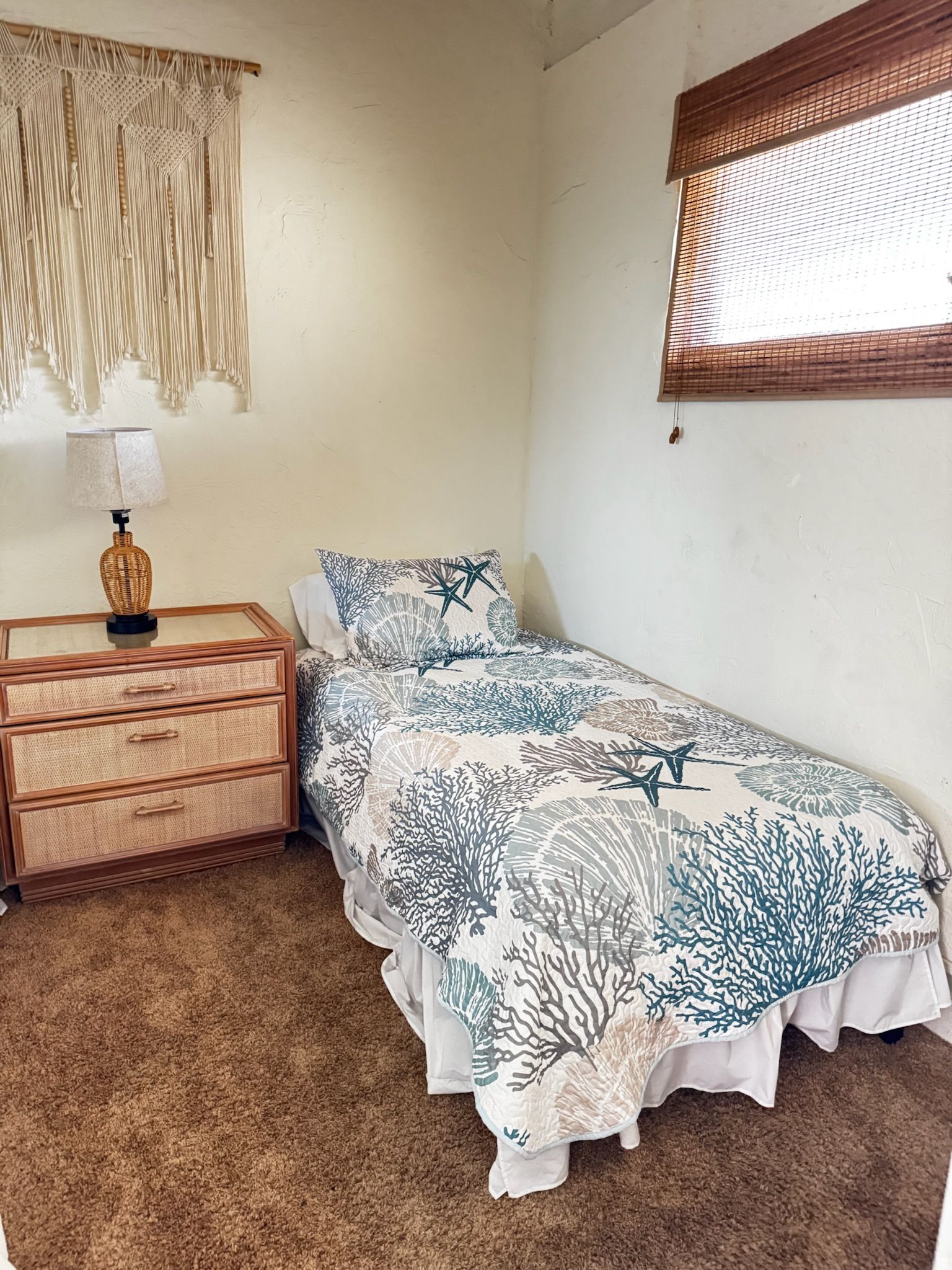 A bedroom with a bed, a nightstand, a lamp, a macrame wall hanging, and a window with blinds.