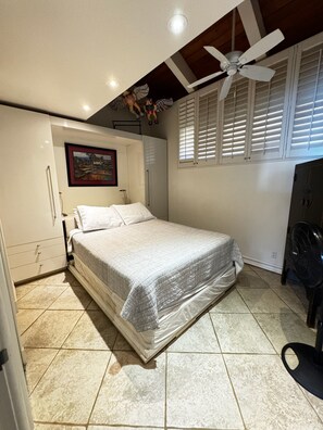 Ein Schlafzimmer mit einem Bett, einem Deckenventilator, einem Fenster mit Jalousien und einem Bild an der Wand.