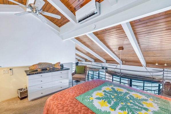 A bedroom with a bed, a dresser, a ceiling fan, and a view of the sea through large windows.