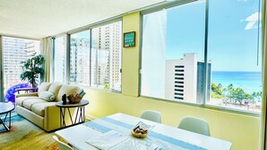 A bright room with a large window, a sofa, a dining table, and a view of the ocean and buildings.