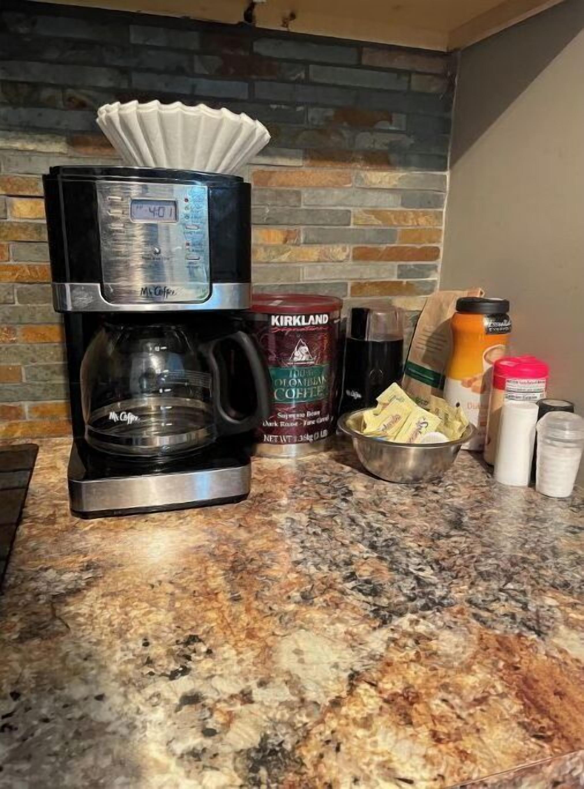 Una cafetera, un recipiente con café Kirkland, un tazón de sobrecitas de azúcar y un frasco de crema sobre una encimera de granito.