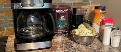 Una cafetera, un recipiente con café Kirkland, un tazón de sobrecitas de azúcar y un frasco de crema sobre una encimera de granito.