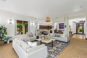 A spacious living area with a sofa set, a coffee table, and a rug. There is a kitchen in the background with wooden cabinets and a bar area with bottles. - Villa Firenze - Peaceful, Private 4BR @PGA (La Quinta)
