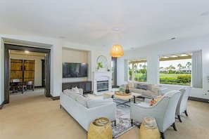 A spacious living room with a fireplace, comfortable seating, a television, and large windows offering a view of a landscaped garden. - Villa Firenze - Peaceful, Private 4BR @PGA (La Quinta)