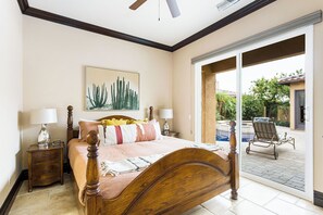 Un dormitorio con una cama grande, dos mesitas de noche, un ventilador de techo y un ventanal corredero que da a un patio exterior con piscina y sillas.
