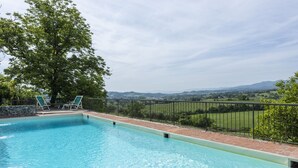 A rectangular swimming pool with clear blue water, surrounded by a black metal fence, and a view of a green landscape with trees and hills in the background. - Villa Poggio Cherici by MMega (Rignano sull'Arno)