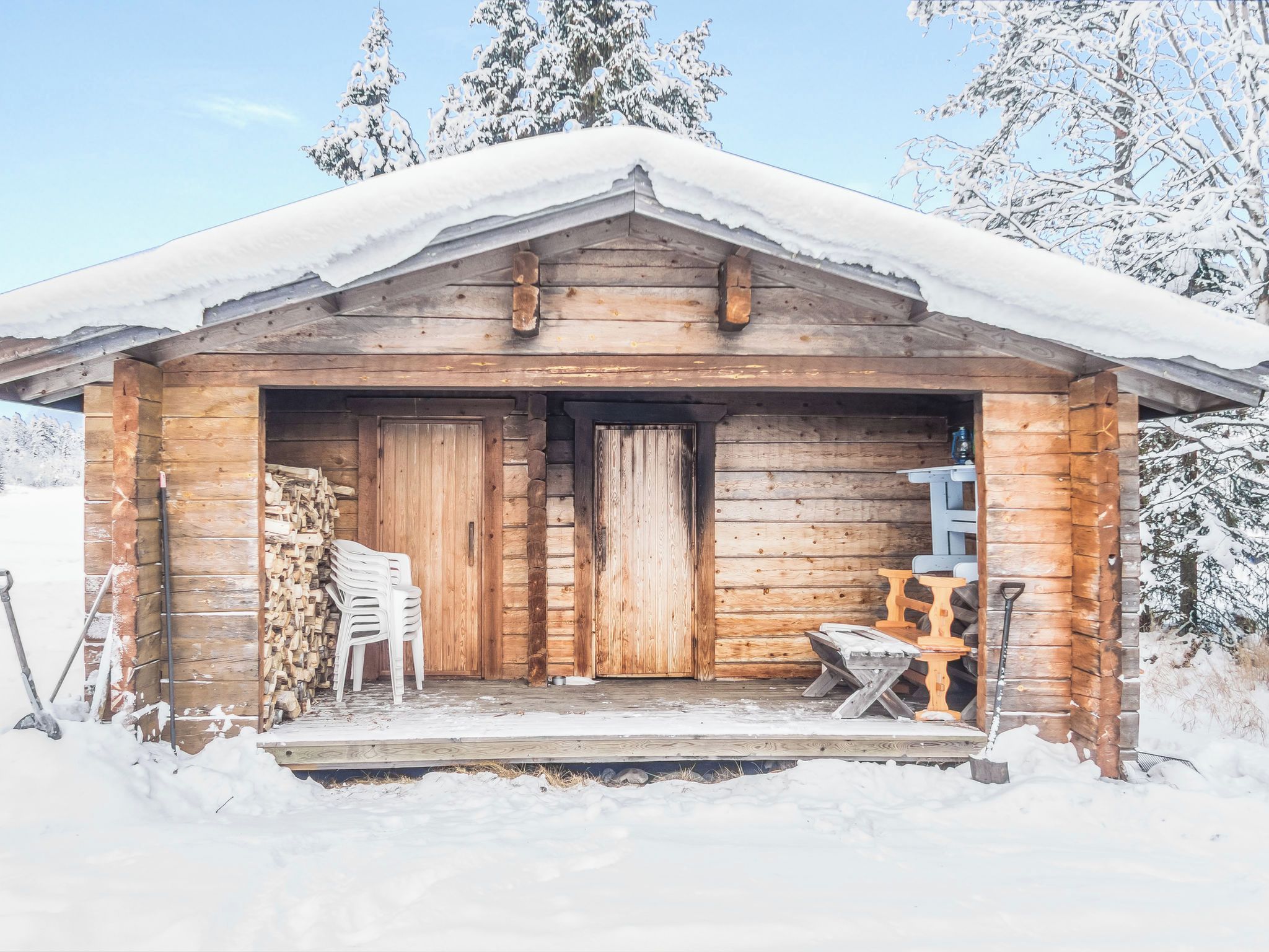 A wooden cabin with a snow-covered roof, a front door, and a small porch area with outdoor furniture.