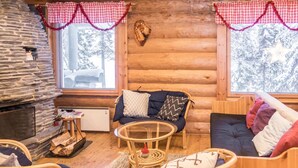 A cozy log cabin interior with a stone fireplace, wicker furniture, and a view of snowy trees through large windows. - Vacation home Mänty by Interhome (Kuusamo)