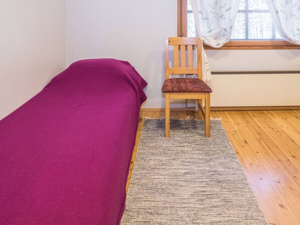 Una cama individual con una colcha roja, una silla de madera con un cojín estampado, y una ventana con cortinas traslúcidas.