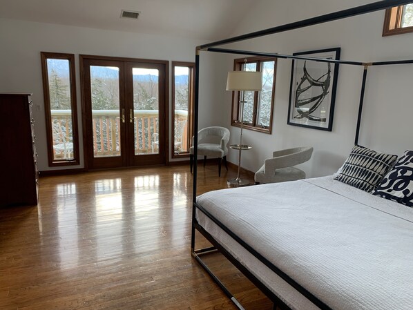 A spacious bedroom with a four-poster bed, a small round table, a chair, and a balcony with a view of trees.