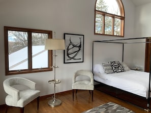 A bedroom with a four-poster bed, two chairs, a floor lamp, and large windows.