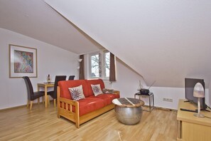 A cozy loft bedroom with a sloped ceiling, a red sofa, a round metal table, and a television on a wooden stand. - Courtyard residence - quiet location and with garden use, close to the sea - Höftresidenz (Middelhagen)