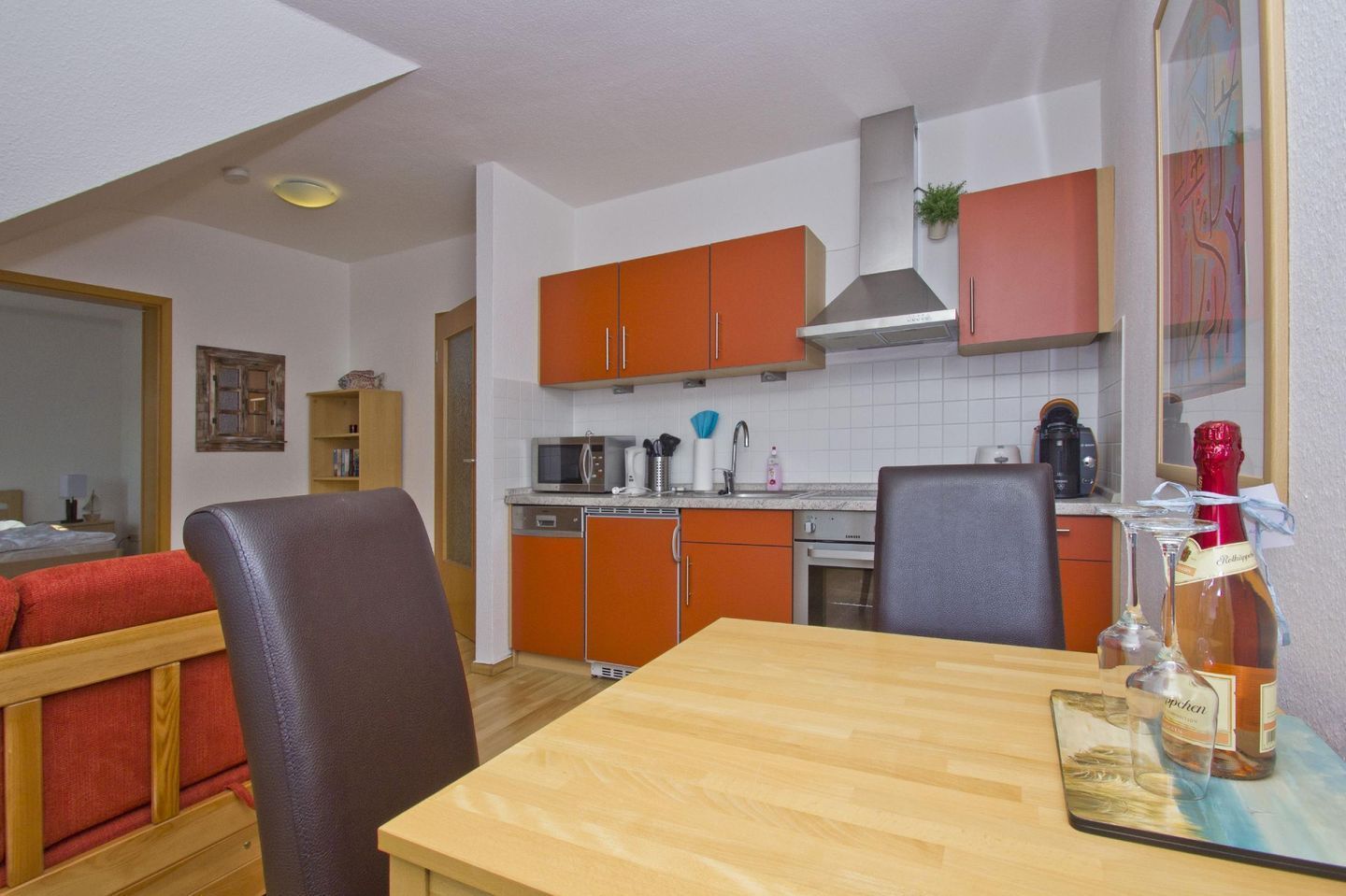 A compact kitchen with orange cabinets, a microwave, a coffee maker, and a bottle of wine on the counter.