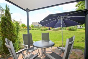 Un patio couvert avec une table et des chaises, un parasol, et une vue sur une pelouse et des bâtiments.