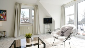 A modern living room with a glass coffee table, a white furry chair, a TV on a stand, and a large window with curtains.