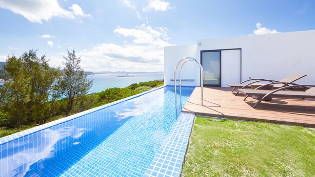 A modern outdoor pool with clear blue water, surrounded by a wooden deck and lounge chairs, overlooking a scenic view of the sea and mountains. - E-horizon Resort Premium SESOKO B (Motobu)