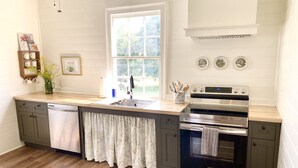 A kitchen with a dishwasher, double sink, stainless steel appliances, wooden flooring, and a window with a view of greenery. - The Jones House: 5 min to VIR, historic 4-bedroom home (Milton)