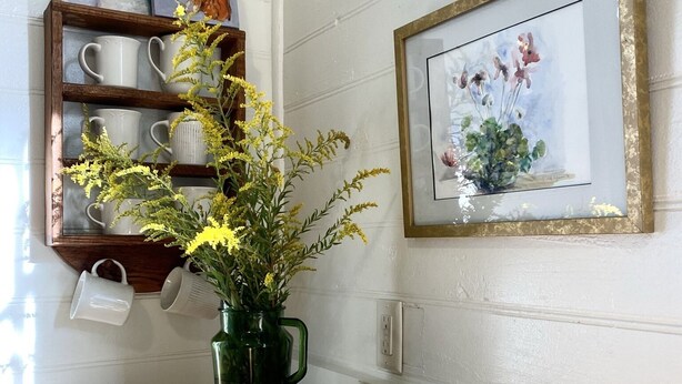 A kitchen counter with a wooden tray holding bread and a vase with flowers. There are mugs on a shelf and a framed painting on the wall. - The Jones House: 5 min to VIR, historic 4-bedroom home (Milton)