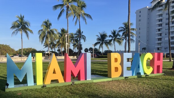 A sign with the words "MIAMI BEACH" in large, colorful letters on a grassy area with palm trees and a building in the background.