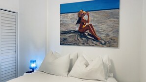 A bedroom with a bed, pillows, a nightstand, a lamp, and a painting of a person on a beach.