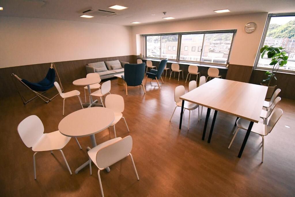 Una sala de reuniones moderna con mesas redondas, sillas blancas, un sofá azul y grandes ventanales con vistas a edificios y zonas verdes.