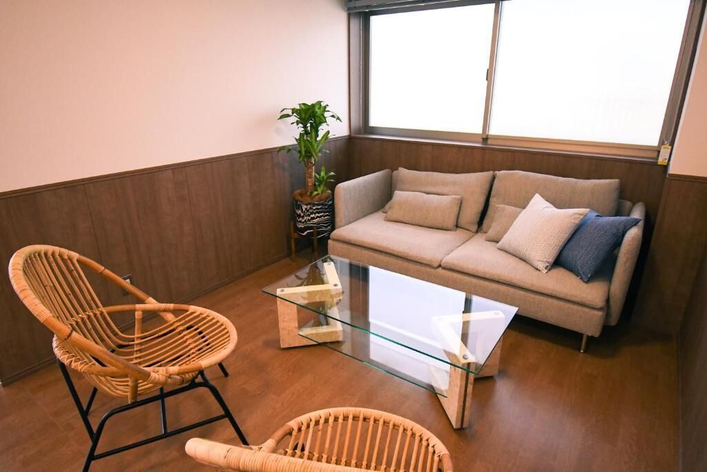 Uma sala de estar com um sofá, duas cadeiras de vime, uma mesa de centro de vidro e um vaso com planta.