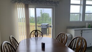 A dining area with a wooden table and chairs, a kitchen with a refrigerator and a dishwasher, and a sliding glass door leading to a deck with a barbecue grill. - Lunker House By the Bay- Rural, Contemporary and Spacious-Moments from Downtown  (Sturgeon Bay)