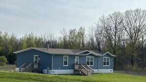 A single-story blue house with a front porch and a small deck, surrounded by a grassy yard and trees. - Lunker House By the Bay- Rural, Contemporary and Spacious-Moments from Downtown  (Sturgeon Bay)