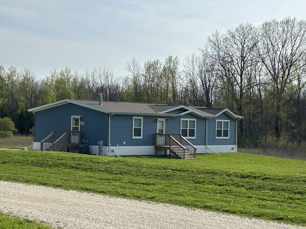 A single-story blue house with a front porch and a small deck, surrounded by a grassy yard and trees.