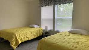 A room with two beds, each with a yellow bedspread and white pillows, a small bedside table with a lamp, and a window with blinds. - Lunker House By the Bay- Rural, Contemporary and Spacious-Moments from Downtown  (Sturgeon Bay)
