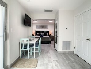 A modern hotel room with a dining area, a bed with a headboard, a mounted TV, and a large window.